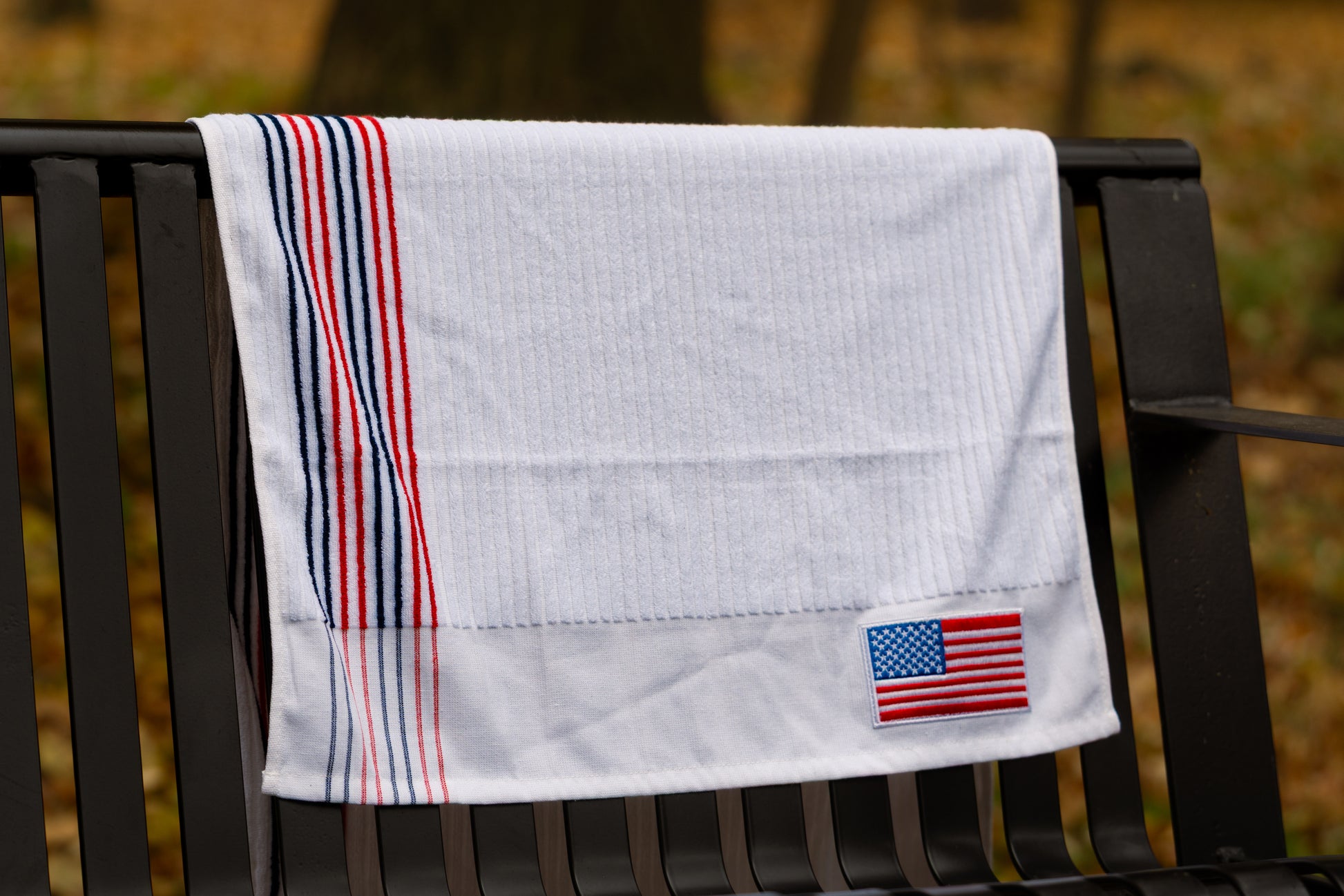 White Pin Hunters Golf Co. looper golf towel draped over a black bench with red and navy stripe detail and American flag patch.