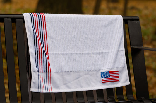 White Pin Hunters Golf Co. looper golf towel draped over a black bench with red and navy stripe detail and American flag patch.
