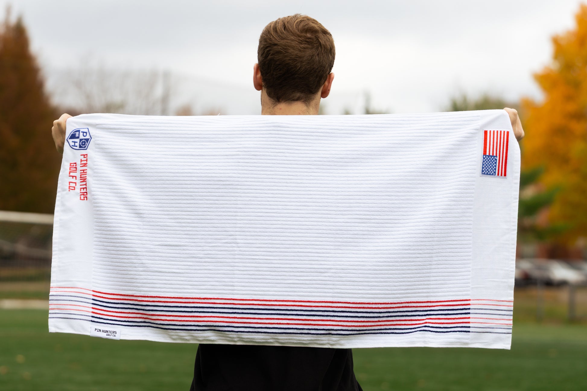 Full view of white Pin Hunters Golf Co. looper golf towel with ribbed cotton texture, red and navy stripe detail, embroidered logo, and American flag patch.