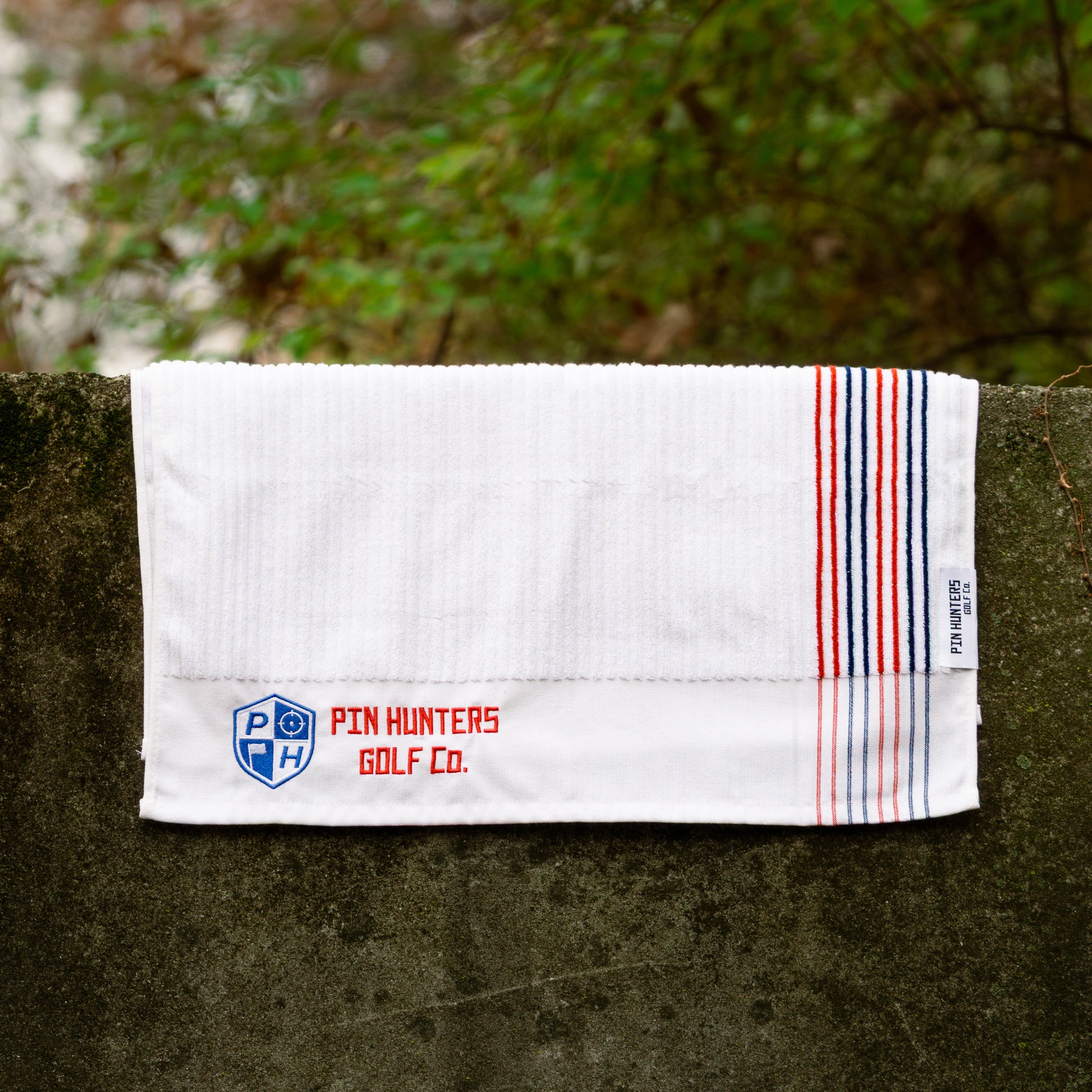White Pin Hunters Golf Co. Looper golf towel displayed on stone with ribbed cotton texture, red and navy stripe detail, and embroidered branding.