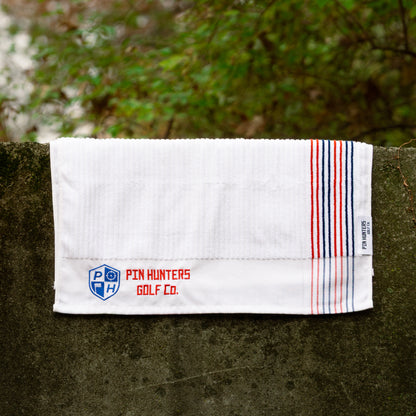 White Pin Hunters Golf Co. Looper golf towel displayed on stone with ribbed cotton texture, red and navy stripe detail, and embroidered branding.