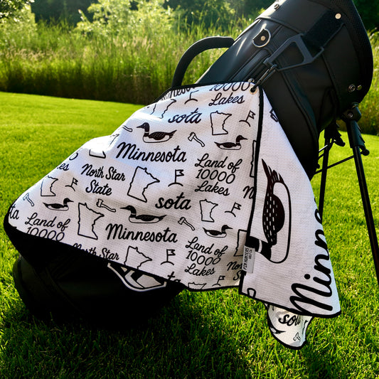 White Minnesota SOTA golf towel hanging from a black golf bag with black loons, Minnesota outlines, North Star State text, and golf icons.