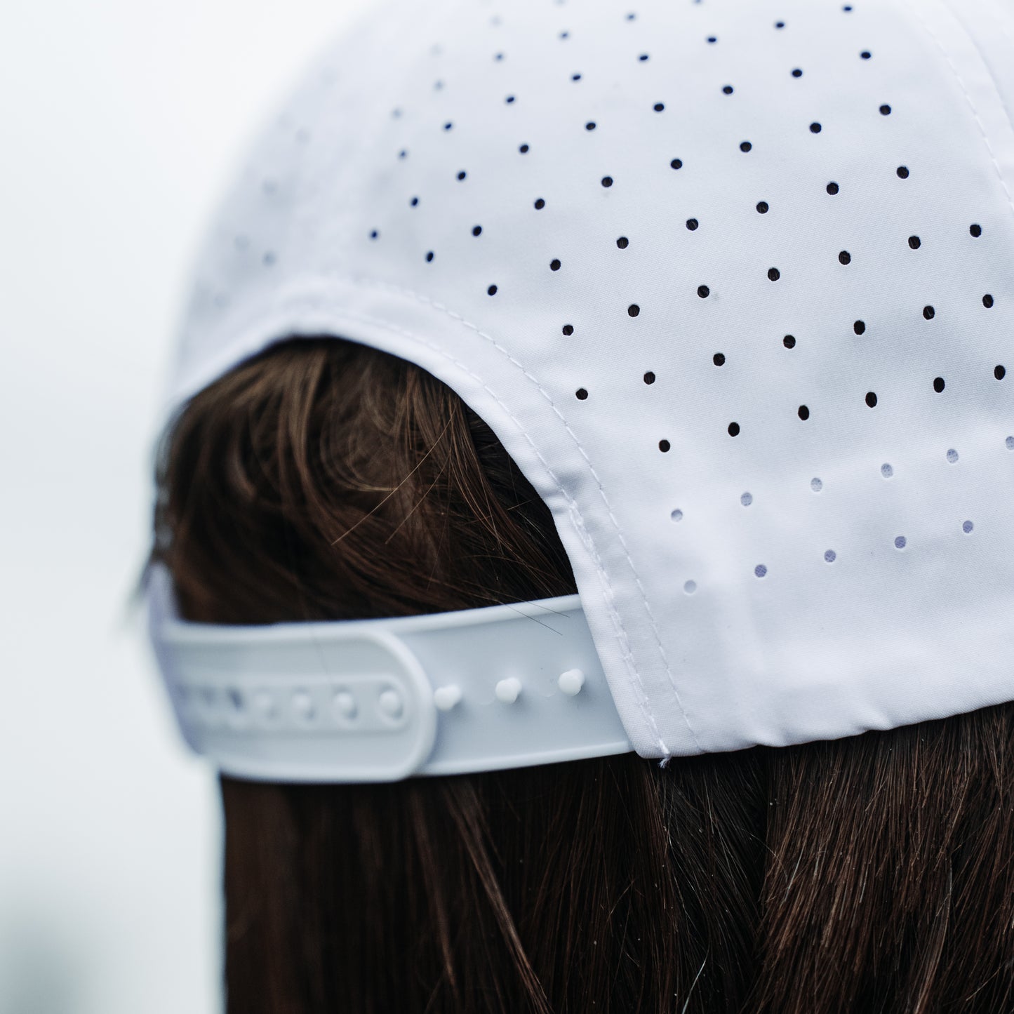 White performance hat back close-up with perforated panels, adjustable snapback closure, curved opening, and clean white stitching.