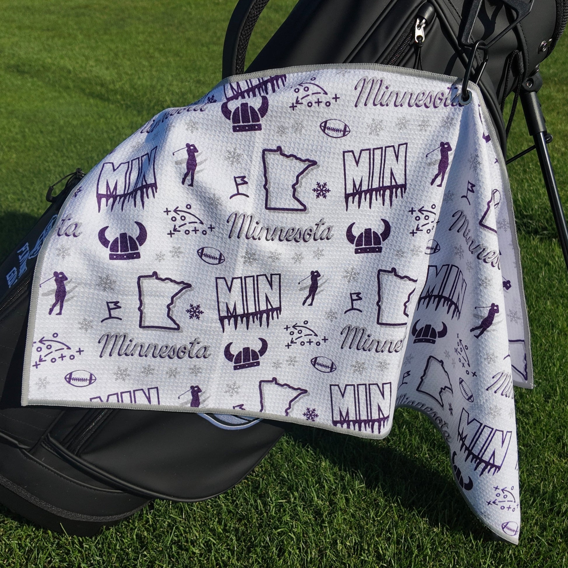 White and purple Minnesota Vikings golf towel hanging on a black golf bag with MIN text, Vikings helmet, football, Minnesota outline, golfer, snowflake, and golf icons.