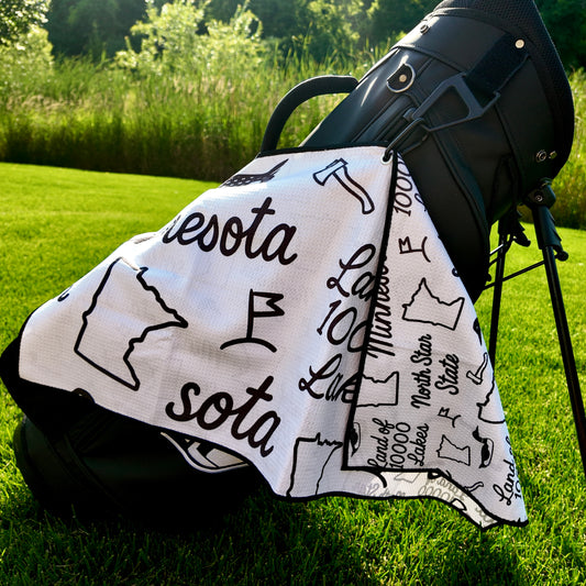 Close up of a white SOTA Minnesota golf towel with black Minnesota outline, loon, Land of 10000 Lakes text, and golf flag graphics.