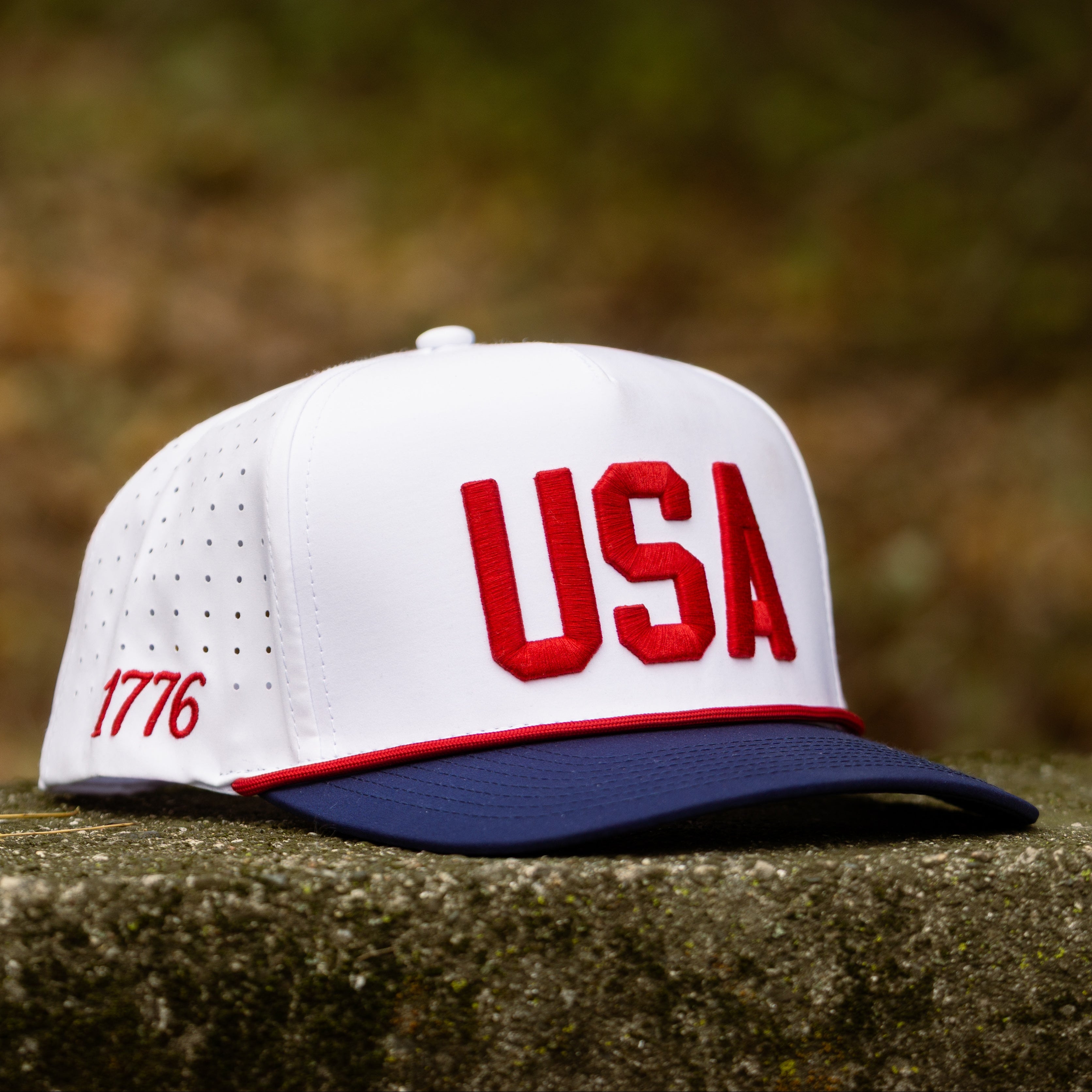 White USA performance golf hat with red raised USA embroidery, navy brim, red rope, and perforated side panels displayed on stone.