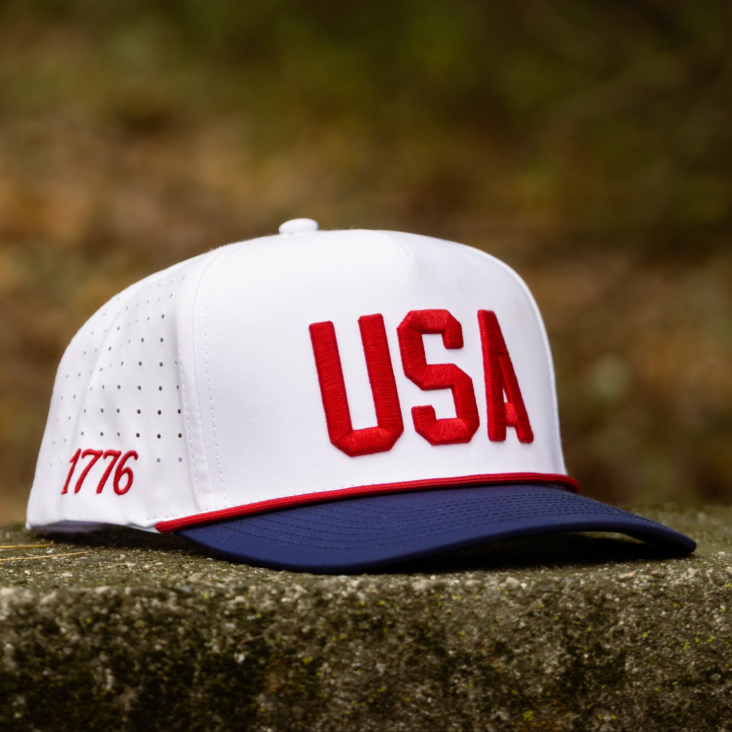White USA performance golf hat with red raised USA embroidery, navy brim, red rope, and perforated side panels displayed on stone.