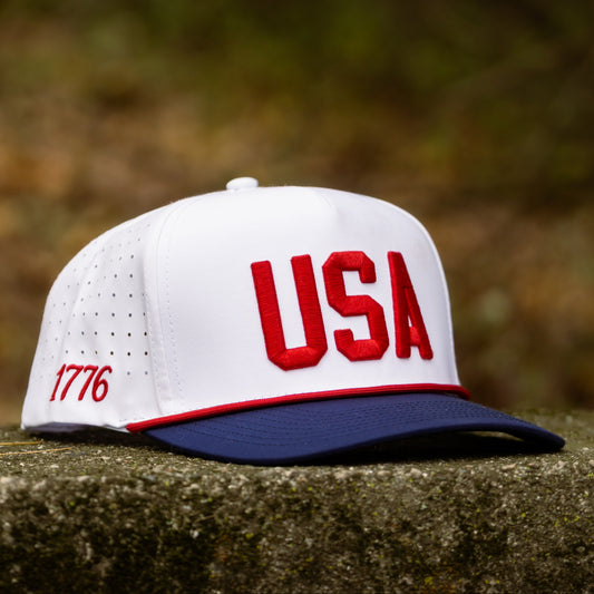 White USA performance golf hat with red raised USA embroidery, navy brim, red rope, and perforated side panels displayed on stone.