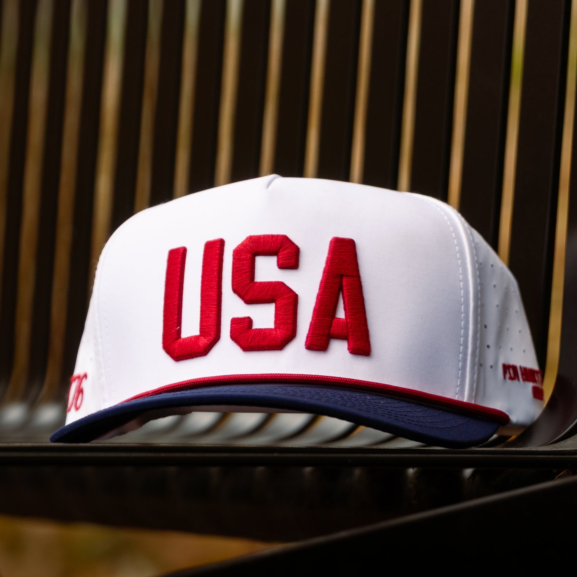 White USA performance golf hat with red raised USA embroidery, red rope, navy brim, and 1776 side embroidery on an outdoor stone background.