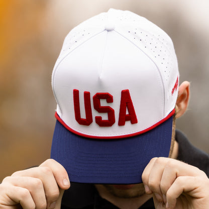 White USA performance golf hat with red raised embroidery, red rope, navy brim, and perforated side panels being held outdoors.