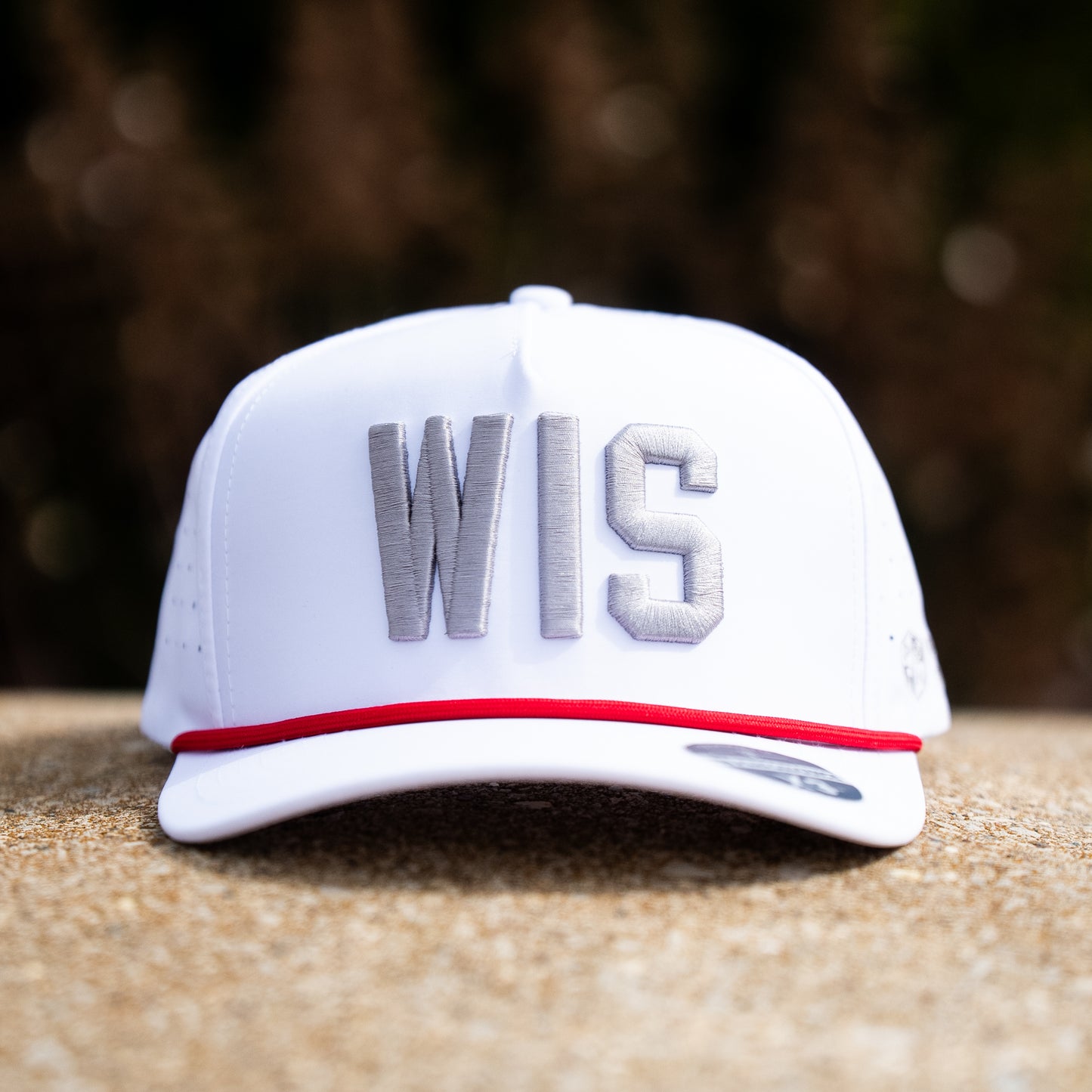 White WIS performance golf hat with gray raised embroidery, red rope, white brim, and clean front view on a stone background.