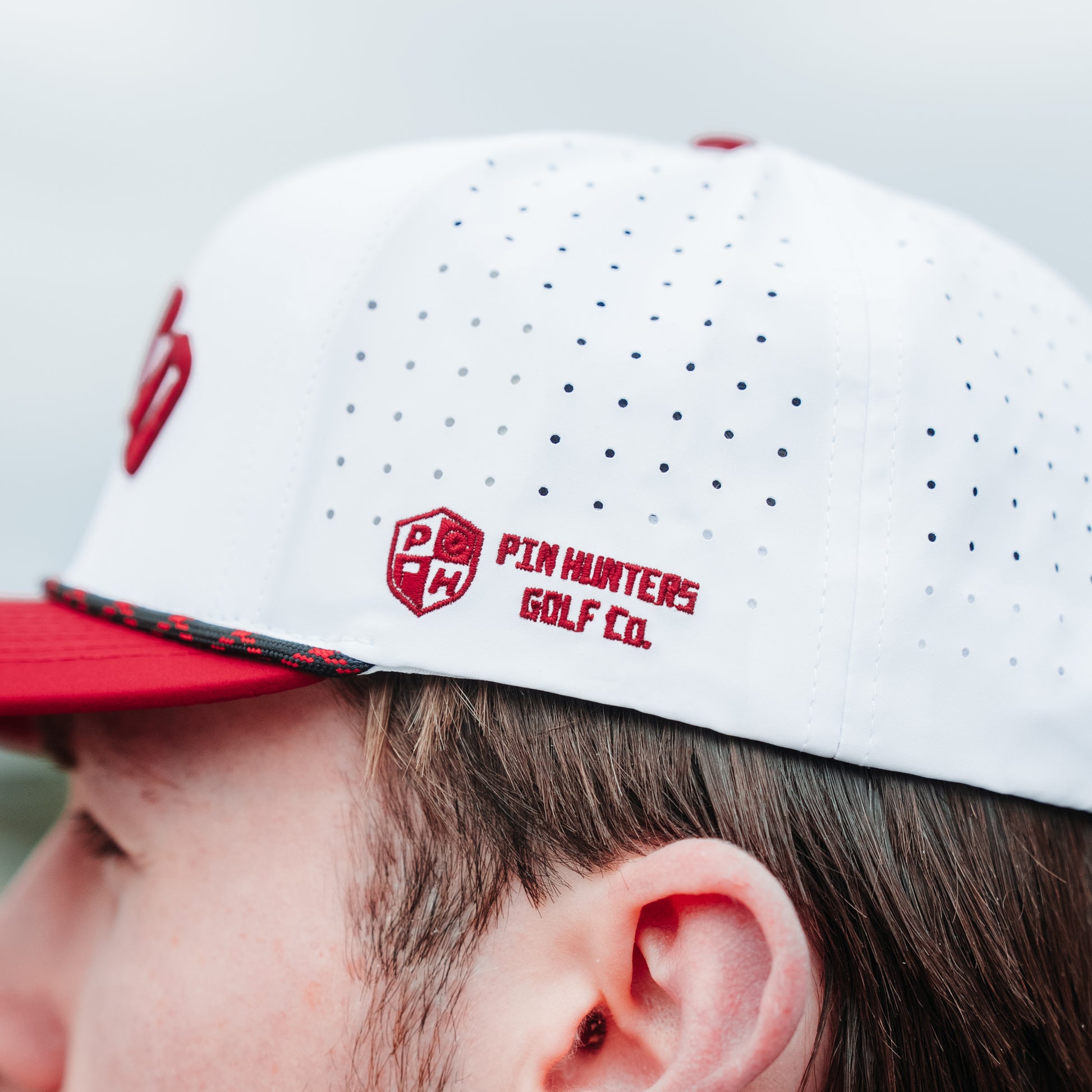 White Wisco performance golf hat side detail with red Pin Hunters logo embroidery, perforated panels, and black and red rope detail.