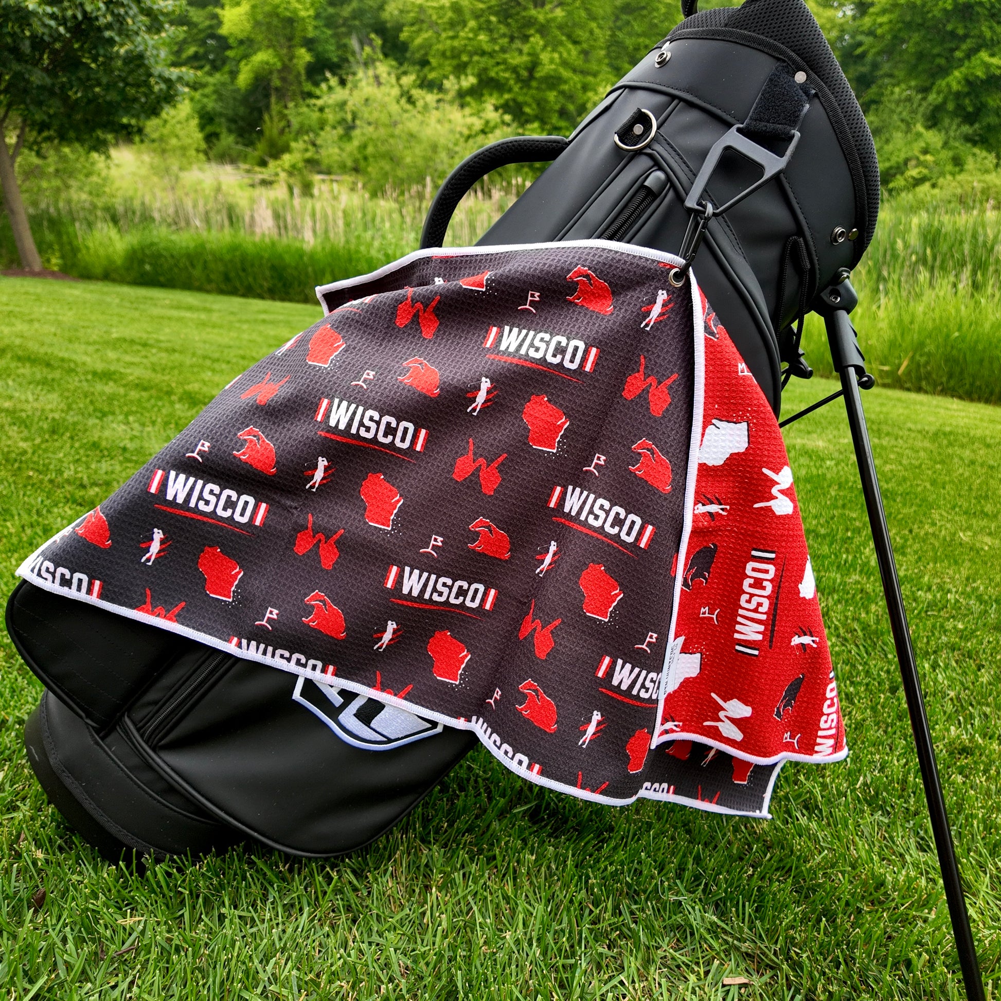 Wisconsin Badgers black golf towel hanging on a black golf bag with I WISCO text, red Wisconsin outlines, badger graphics, golfers, golf flags, and red reverse side.